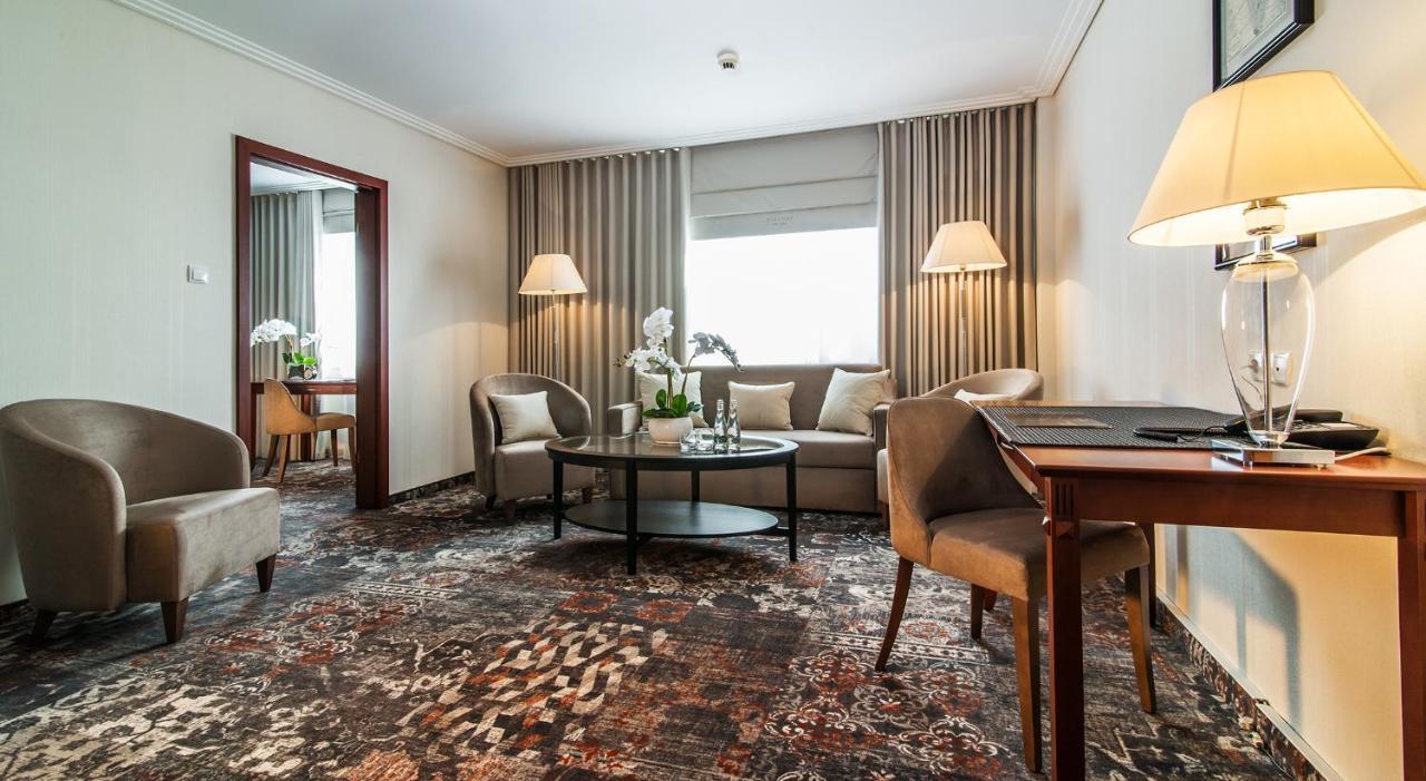 エアポート ホテル オケンチェ ワルシャワ エクステリア 写真 The photo shows a well-furnished hotel room or lounge area. It features a seating arrangement with two armchairs and a sofa, all positioned around a central coffee table. There are also two lamps providing ambient lighting. The decor includes light-c